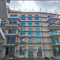 Budynek wielorodzinny w trakcie remontu elewacji, okna zabezpieczone folią, widoczne rusztowania.