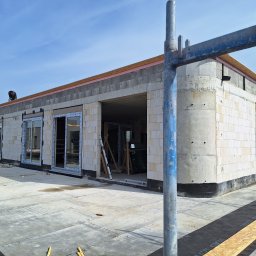 Bartosz Doszko... - Budynek w trakcie budowy z zamontowanymi oknami i drzwiami przesuwnymi, widoczny pracownik na dachu, rusztowanie w pierwszym planie.