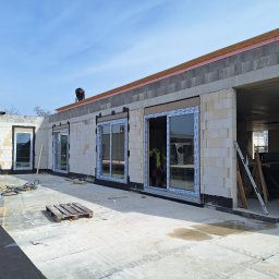 Bartosz Doszko... - Budynek w trakcie budowy z zamontowanymi oknami balkonowymi, widoczne materiały budowlane i narzędzia, pracownik na dachu