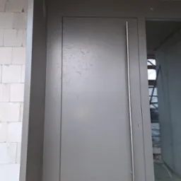 Szare drzwi zewnętrzne z długim, pionowym uchwytem, zamontowane w surowym stanie budynku, widoczne ślady po montażu i pianka uszczelniająca.