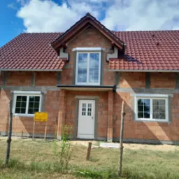 Surowy stan domu jednorodzinnego z czerwonej cegły z zamontowanymi białymi oknami, białą stolarką okienną i drzwiami wejściowymi, widoczne rusztowanie i tablica informacyjna.