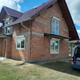 Dom w Zgorzelcu w trakcie budowy, widoczne białe okna z częściowo opuszczonymi roletami zewnętrznymi i surowa elewacja z cegły.