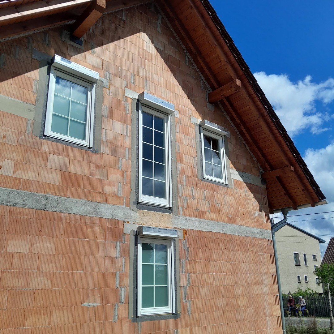 Surowy mur z cegły z zamontowanymi białymi oknami z roletami zewnętrznymi w trakcie montażu, widok z boku na tle błękitnego nieba.