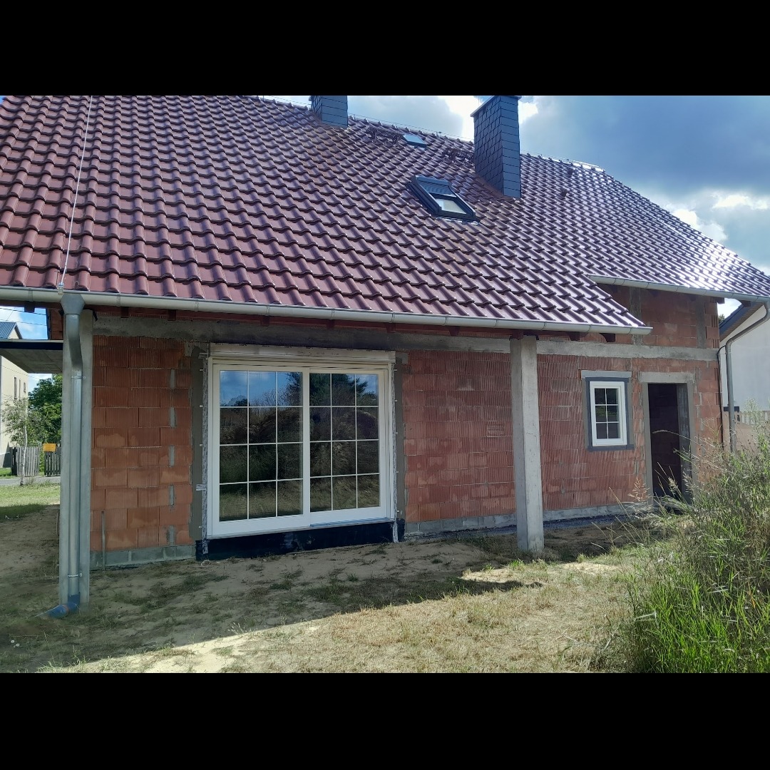 Nowy dom w trakcie budowy z zamontowanymi białymi oknami PCV z podziałem na szprosy, widoczne cegły i dach z czerwoną dachówką.