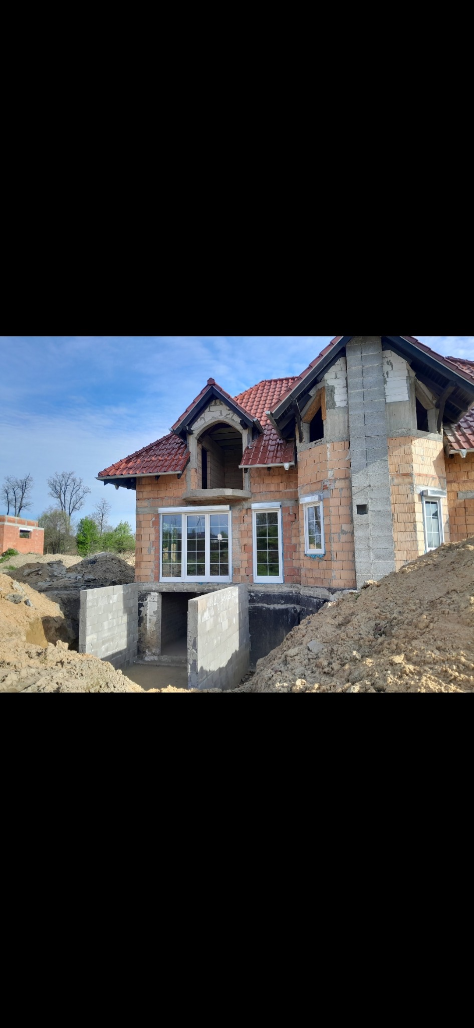 Budynek w trakcie budowy z zamontowanymi białymi oknami, widoczne fundamenty i ceglana elewacja, teren wokół rozkopany.