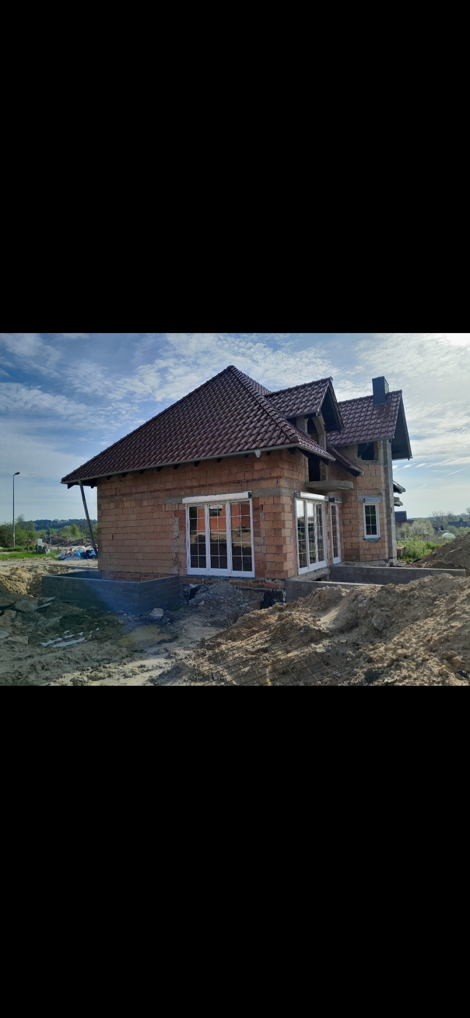 Dom w trakcie budowy z zamontowanymi białymi oknami i drzwiami balkonowymi, widoczne cegły i czerwona dachówka, teren wokół domu w trakcie prac ziemnych.