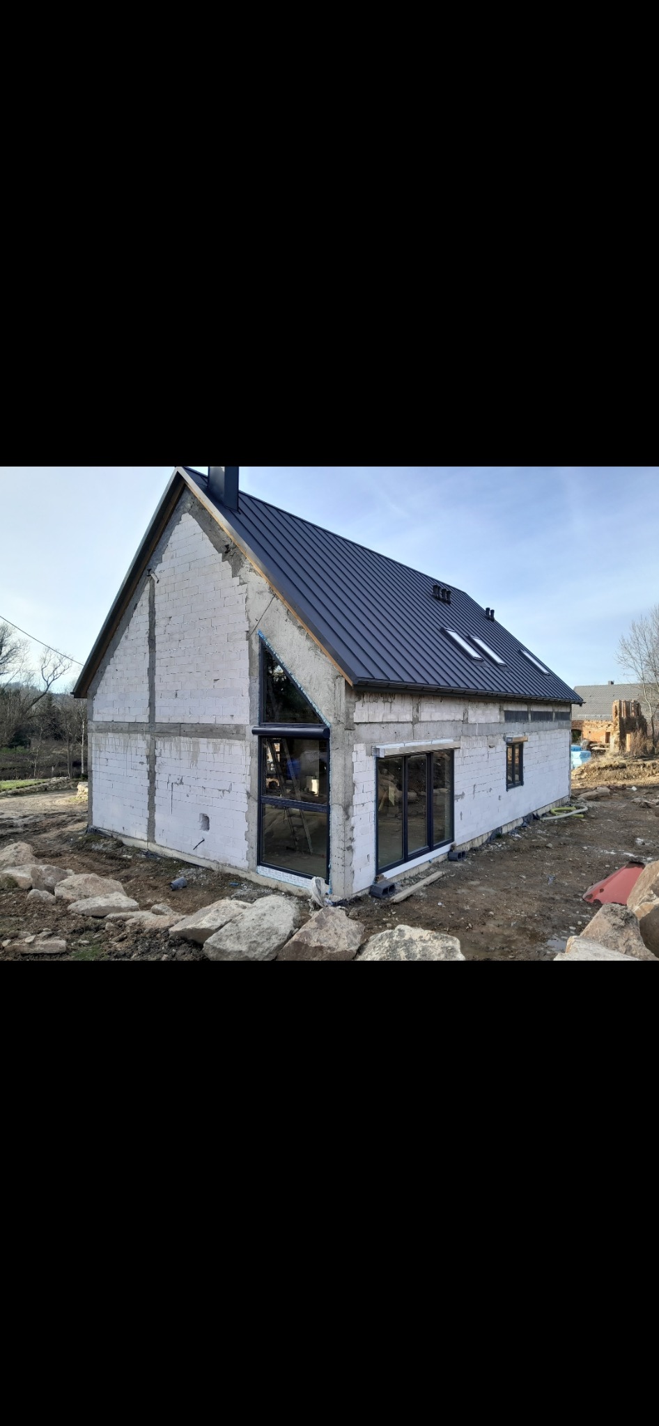 Surowy stan budynku z oknami w czarnych ramach i roletą zewnętrzną częściowo opuszczoną na trójkątnym oknie, widoczne rusztowania wewnątrz, czarny dach i gruz przed budynkiem.