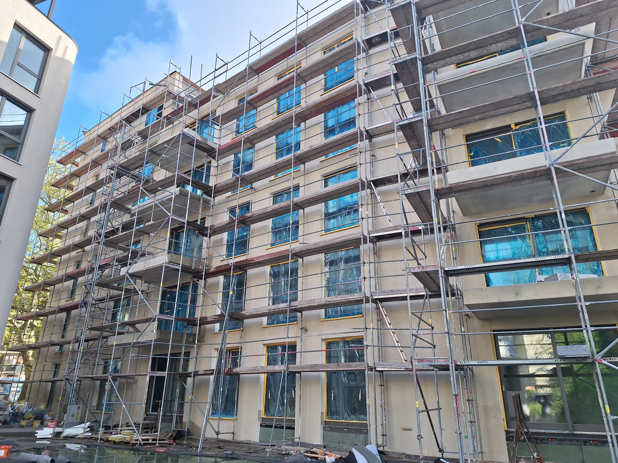 Budynek w trakcie prac elewacyjnych, okna zabezpieczone folią, rusztowania na całej fasadzie, widok z dołu na kilka pięter.