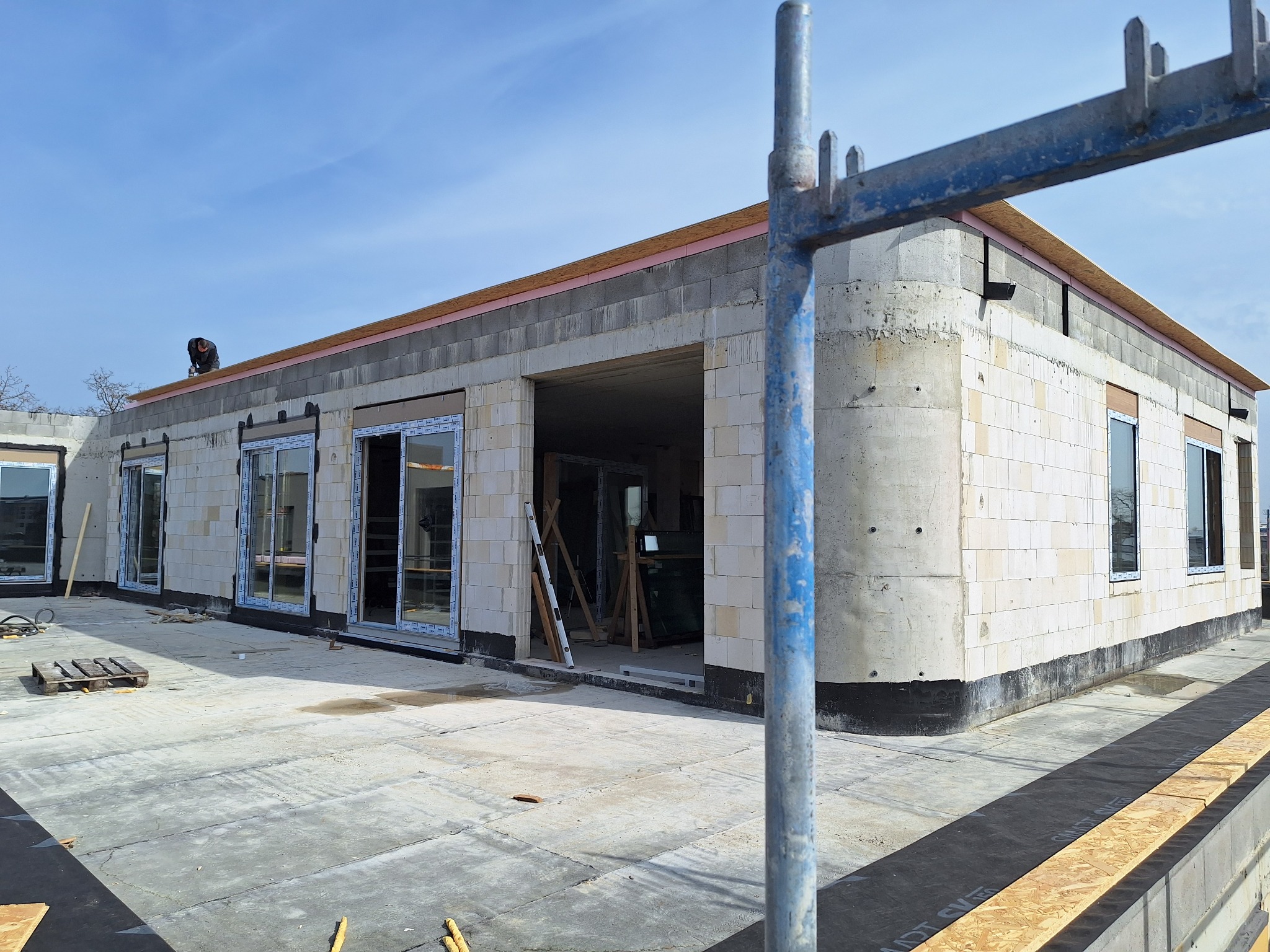 Budynek w trakcie budowy z zamontowanymi oknami i drzwiami przesuwnymi, widoczny pracownik na dachu, rusztowanie w pierwszym planie.