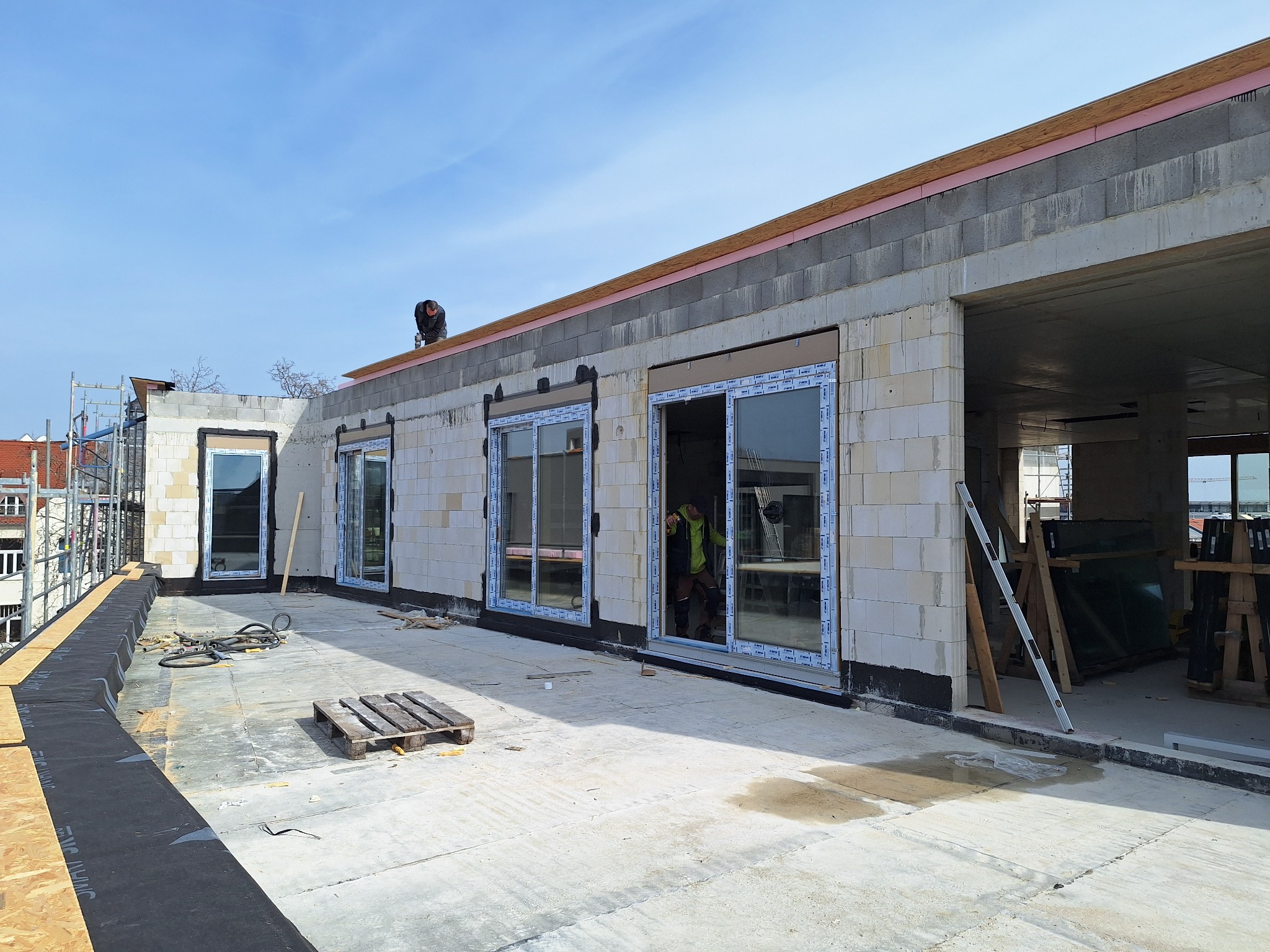 Budynek w trakcie budowy z zamontowanymi oknami balkonowymi, widoczne materiały budowlane i narzędzia, pracownik na dachu