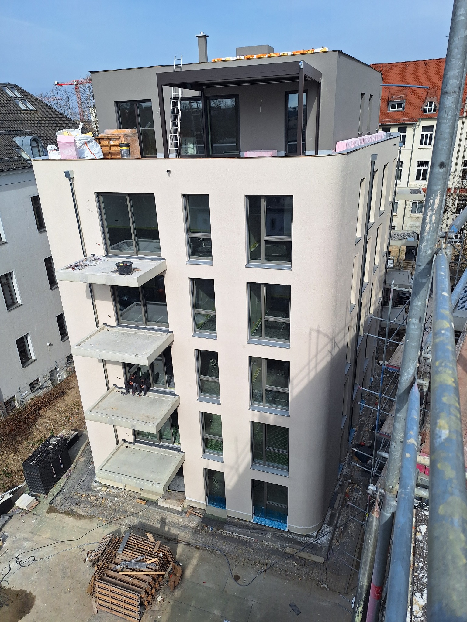 Wysoki budynek mieszkalny z balkonami i oknami PCV w trakcie montażu, widok z góry z rusztowania, na balkonach robotnicy w ciemnych ubraniach.