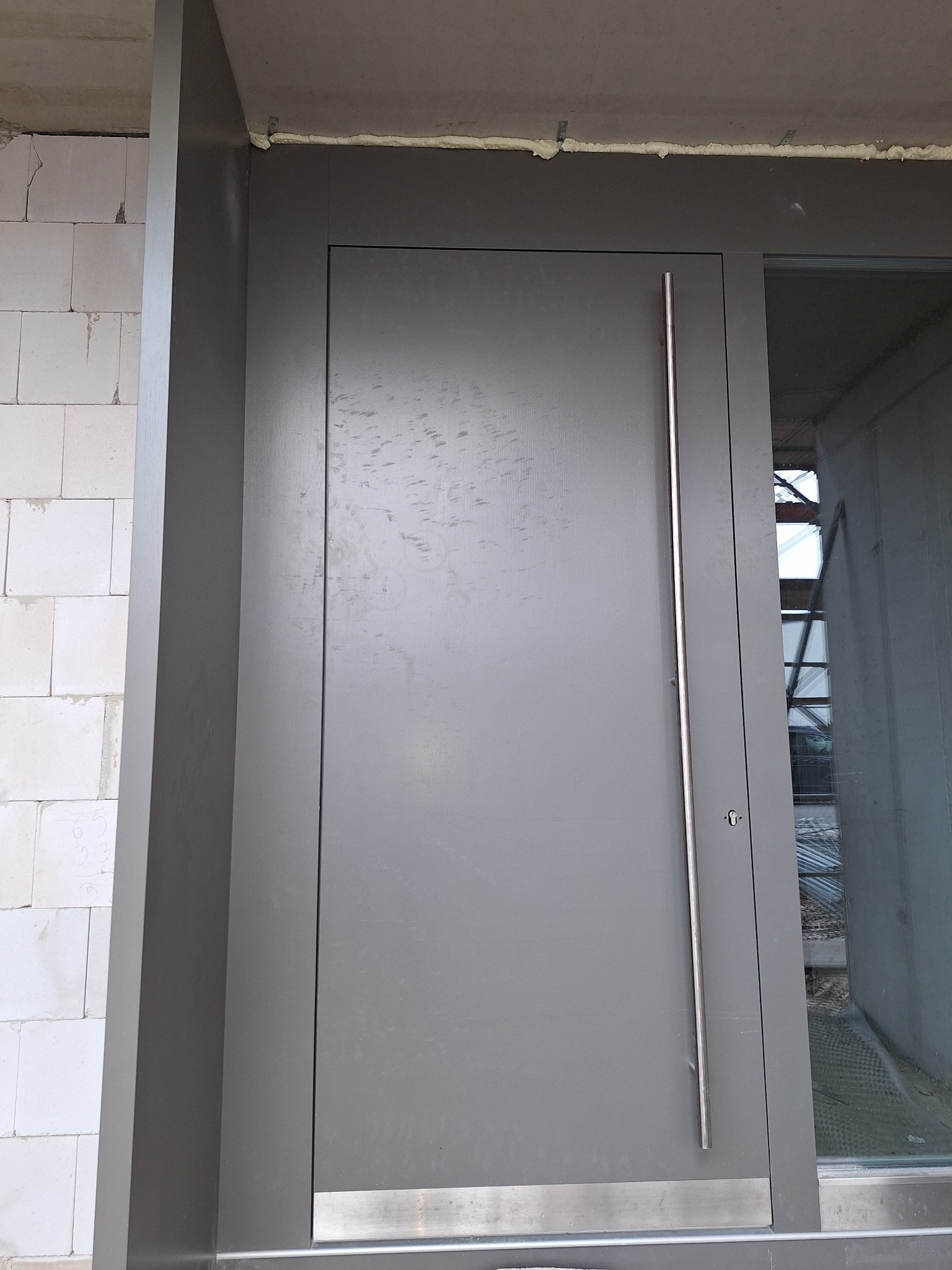 Szare drzwi zewnętrzne z długim, pionowym uchwytem, zamontowane w surowym stanie budynku, widoczne ślady po montażu i pianka uszczelniająca.