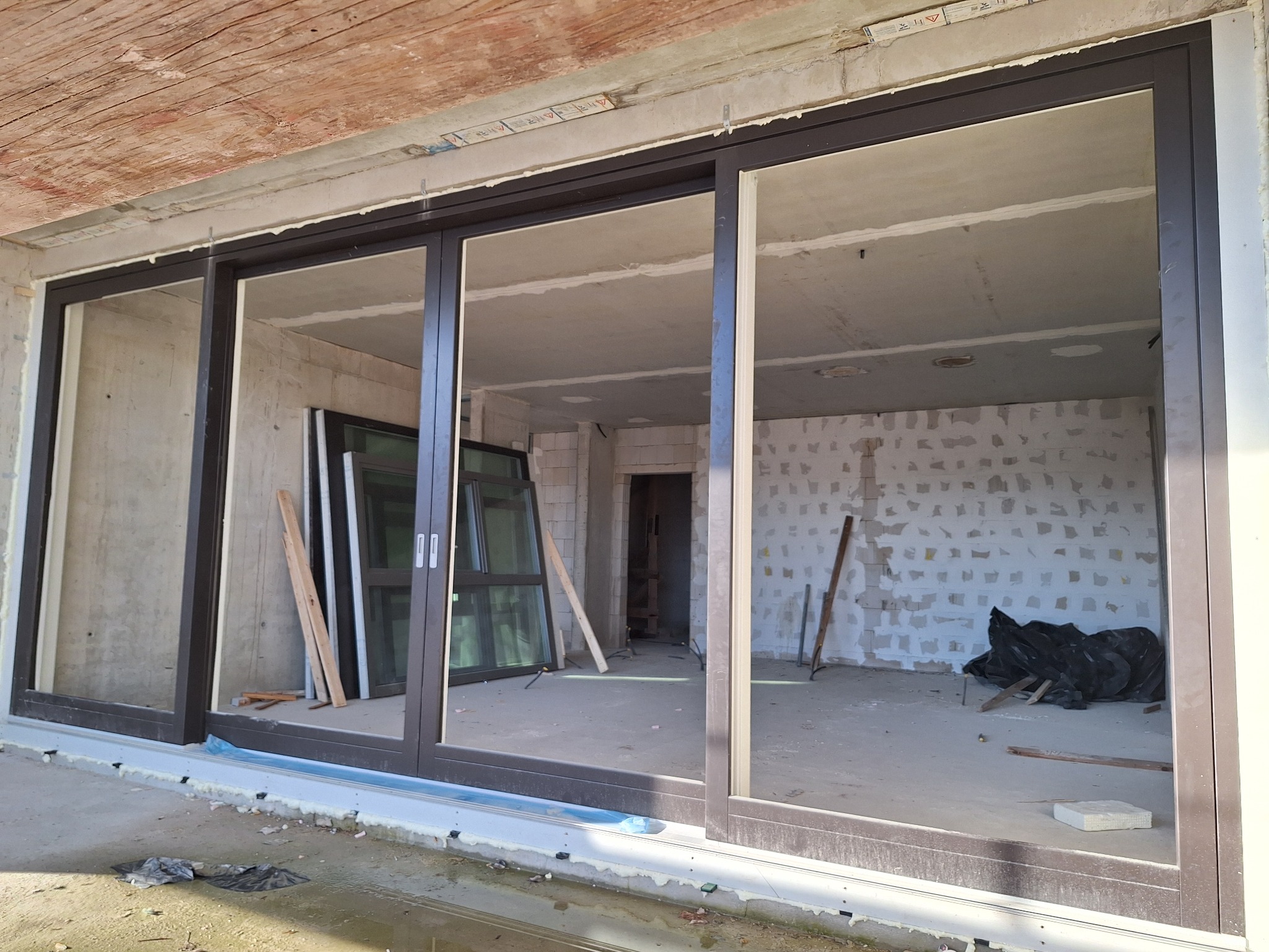 Montaż dużych, brązowych drzwi przesuwnych w surowym stanie budynku, widoczne materiały budowlane i pianka montażowa.