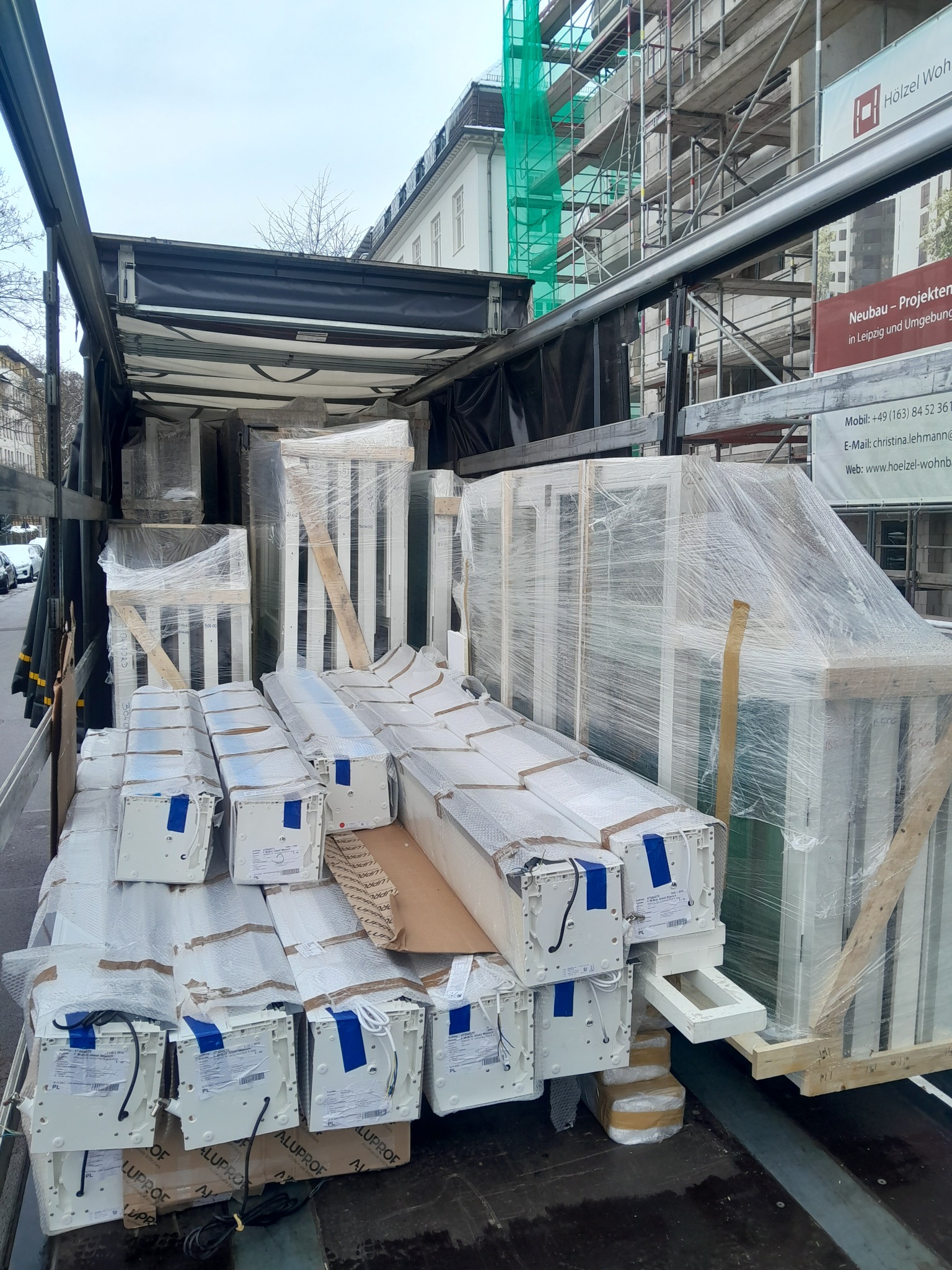 Transport okien i rolet zapakowanych w folię na pace ciężarówki, widoczne skrzynie z roletami z napisem Aluprof, w tle budynek w rusztowaniach.