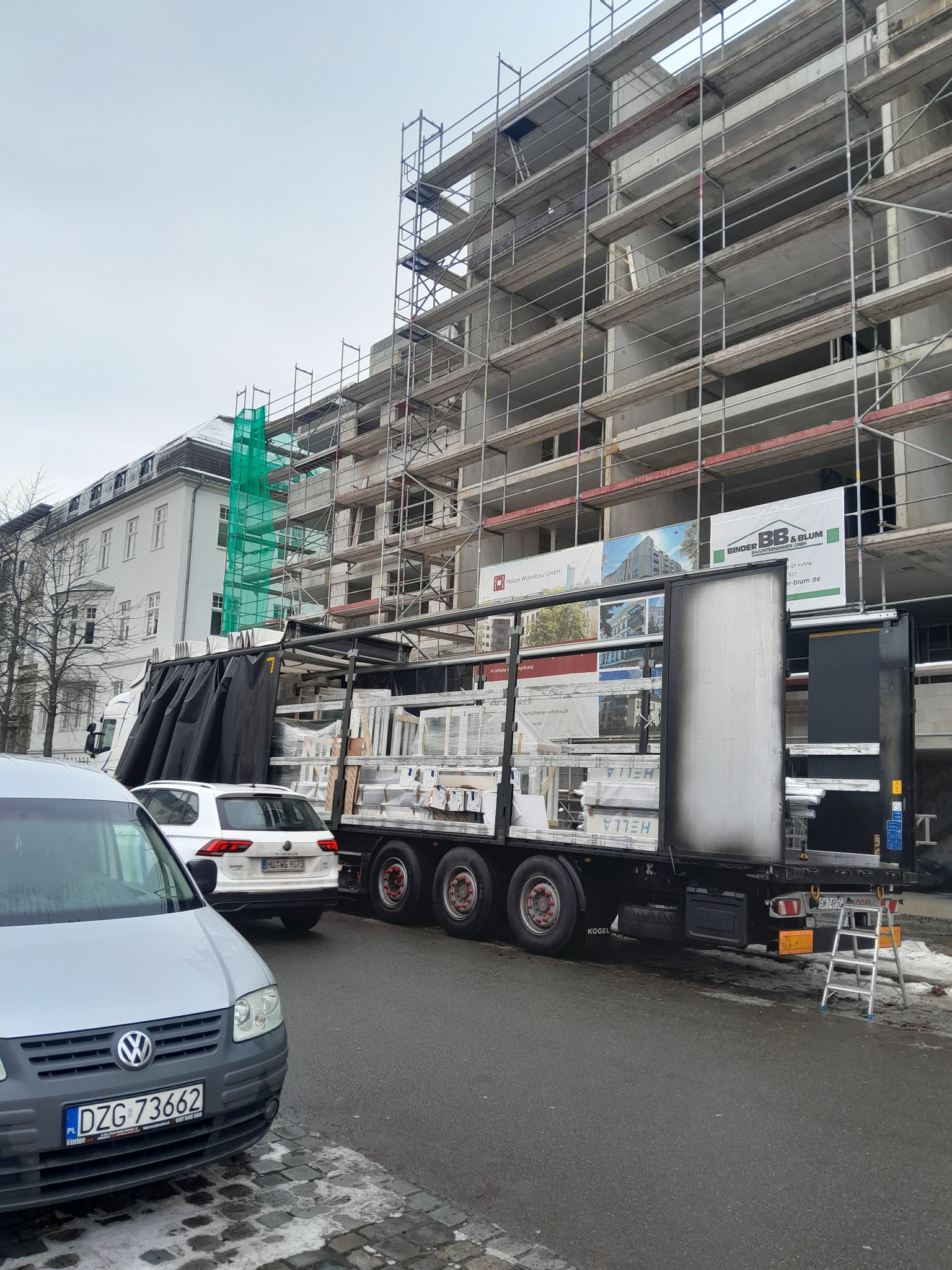 Ciężarówka z ładunkiem okien PCV marki HELLA zaparkowana na ulicy przed budynkiem w trakcie budowy, z widocznymi rusztowaniami i reklamami firm budowlanych.