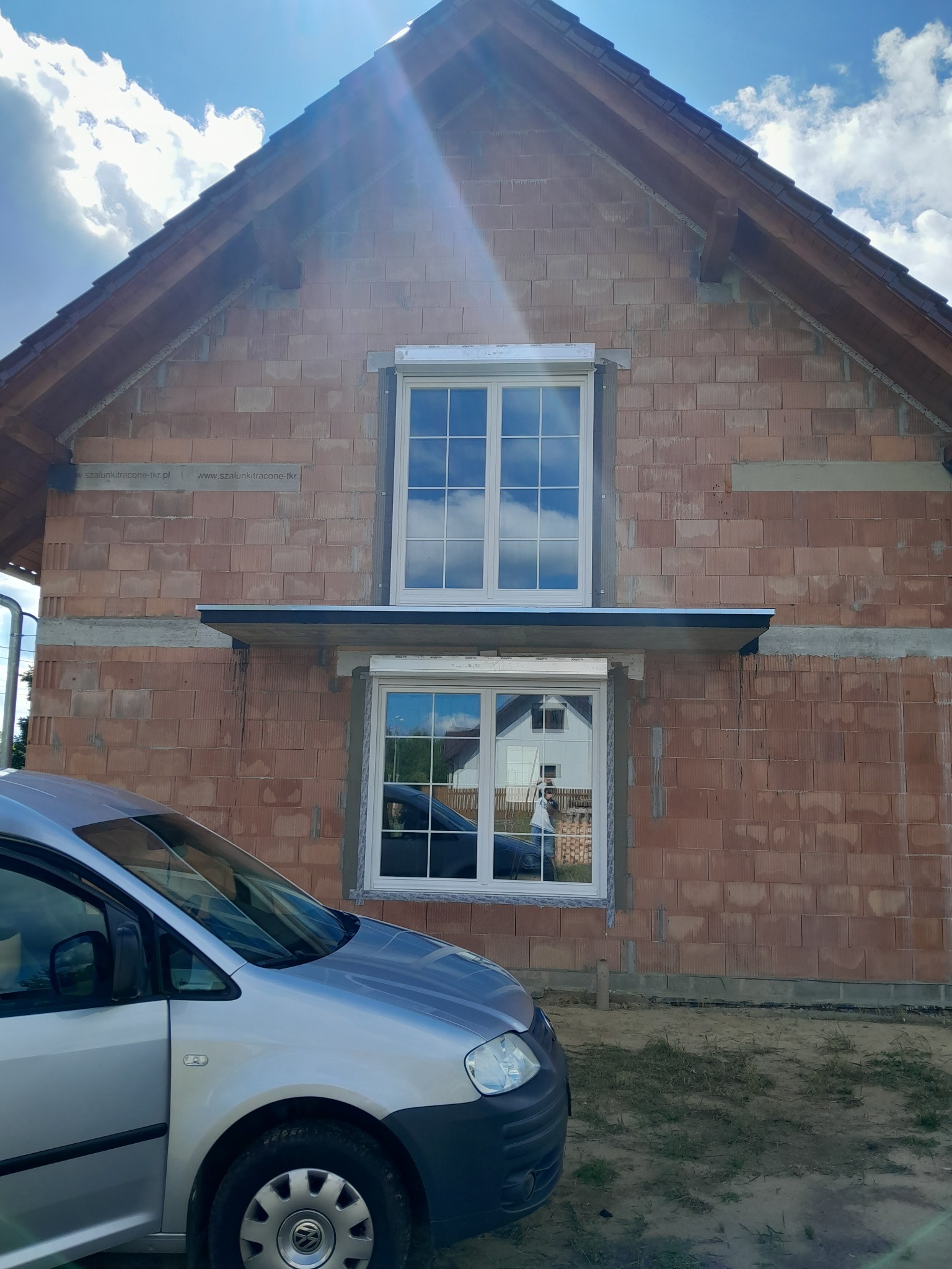 Nowe, białe okna z roletą zewnętrzną zamontowane w surowym budynku z cegły, widoczne odbicie w szybie, samochód dostawczy na pierwszym planie.