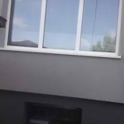 Szara elewacja domu z białymi oknami i małym oknem piwnicznym, widoczne odbicie nieba w szybach.