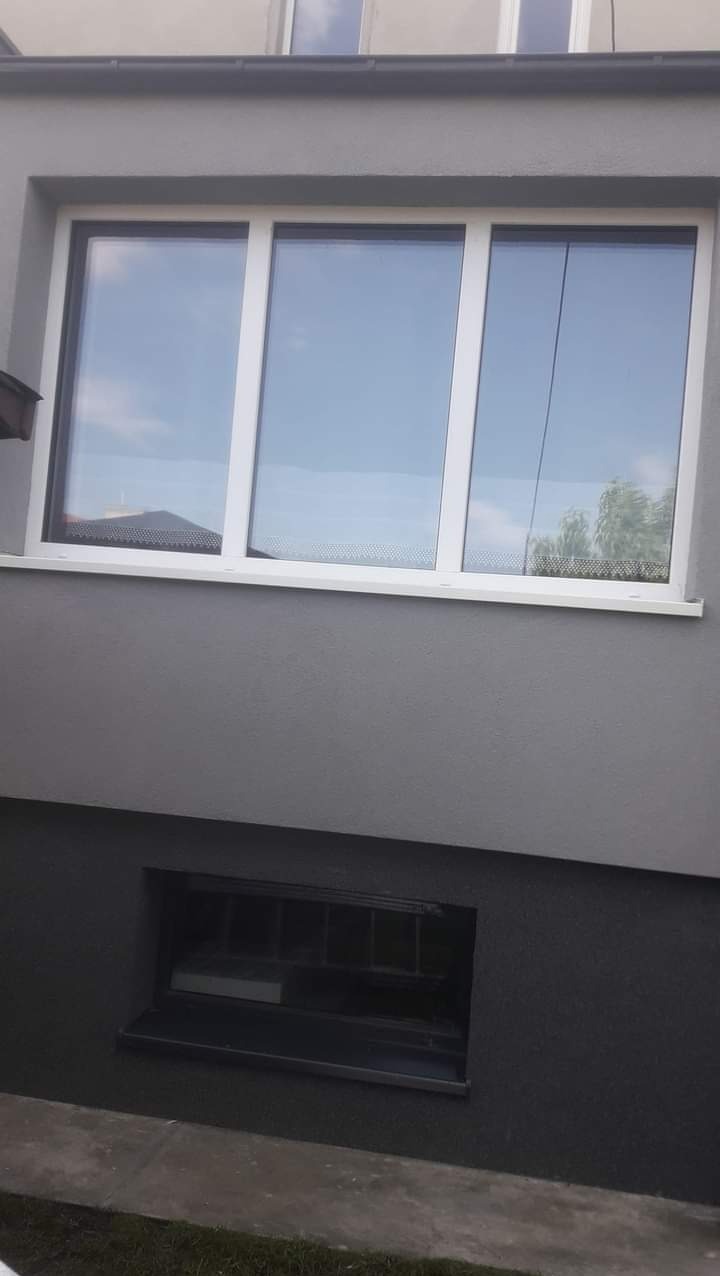 Szara elewacja domu z białymi oknami i małym oknem piwnicznym, widoczne odbicie nieba w szybach.