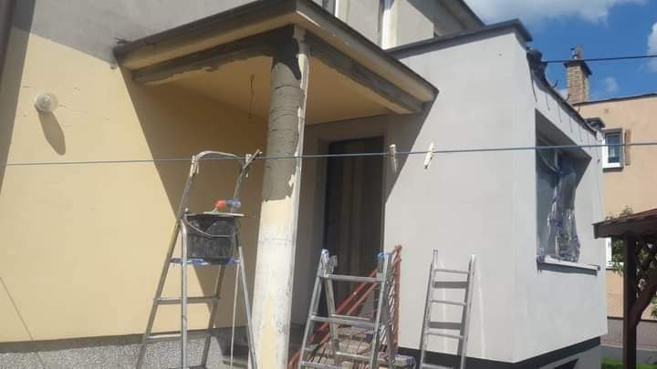 Elewacja domu w trakcie malowania; widoczne rusztowania, wiadro z farbą i częściowo pomalowana kolumna wejściowa.