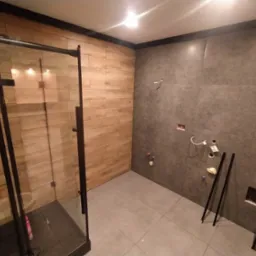 Wnętrze łazienki w trakcie wykańczania: kabina prysznicowa z czarnymi profilami, drewnopodobne płytki na jednej ścianie, szare płytki imitujące beton na drugiej, widoczne instalacje hydrauliczne...