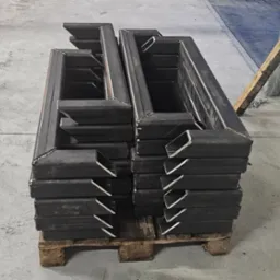 Stos spawanych, metalowych profili zamkniętych o przekroju kwadratowym, ułożonych na palecie w hali produkcyjnej. Widoczny przewód zasilający i brama garażowa w tle.