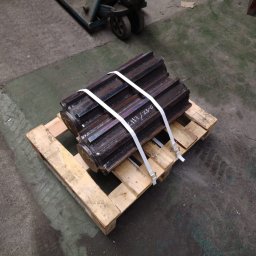 LEGAL & TECHNICAL SUPPORT CENTRE SPÓŁKA Z OGRANICZONĄ ODPOWIEDZIALNOŚCIĄ - Dwa duże, metalowe koła zębate, zabezpieczone białymi pasami, leżące na drewnianej palecie na tle betonowej posadzki warsztatu. Widoczne ślady obróbki i napisy na powierzchni metalu.