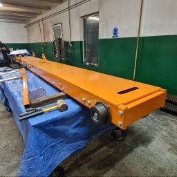 LEGAL & TECHNICAL SUPPORT CENTRE SPÓŁKA Z OGRANICZONĄ ODPOWIEDZIALNOŚCIĄ - Pomarańczowa konstrukcja stalowa na wózku warsztatowym, widoczne mocowania śrubowe, koła, w tle butla z gazem i pracownik przeglądający telefon w warsztacie.