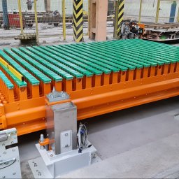 LEGAL & TECHNICAL SUPPORT CENTRE SPÓŁKA Z OGRANICZONĄ ODPOWIEDZIALNOŚCIĄ - Pomarańczowa konstrukcja z zielonymi elementami, prawdopodobnie część większej maszyny, ustawiona na szarym betonowym podłożu w hali produkcyjnej. Widoczne elementy automatyki i czujniki.