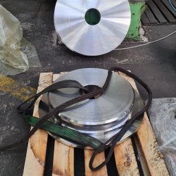 LEGAL & TECHNICAL SUPPORT CENTRE SPÓŁKA Z OGRANICZONĄ ODPOWIEDZIALNOŚCIĄ - Srebrne, okrągłe elementy z centralnym otworem, ułożone na drewnianej palecie z pasami transportowymi, w tle widoczna zielona tokarka.