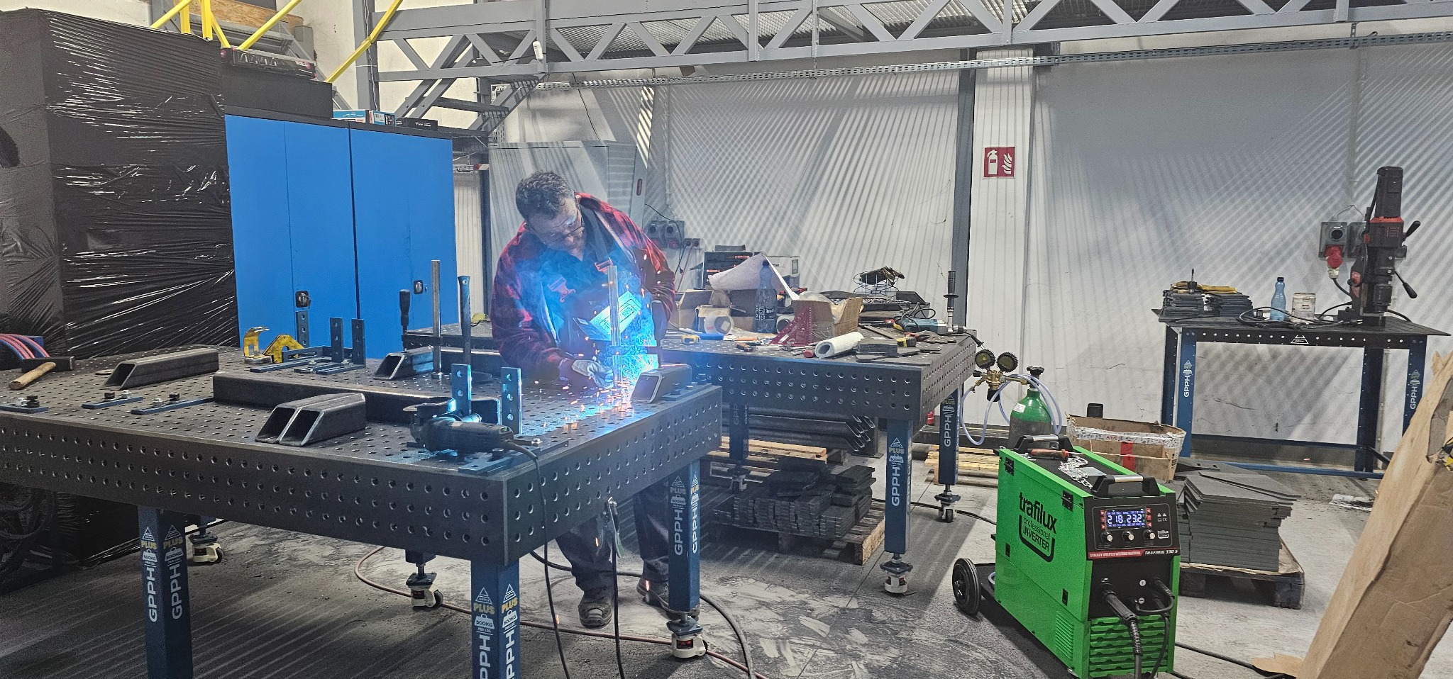 Spawanie elementu na perforowanym stole spawalniczym. Widoczne iskry i spawacz w masce ochronnej, używający sprzętu Trafilux. W tle warsztat z niebieskimi szafkami i konstrukcją stalową pod sufitem.