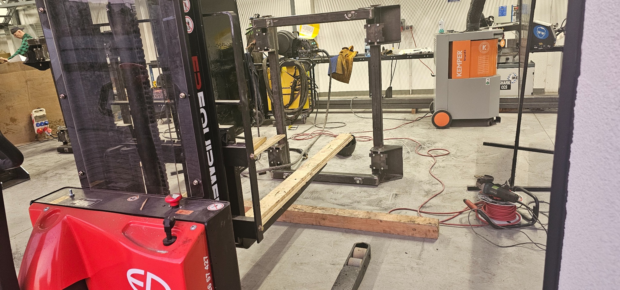 Wózek widłowy z zamontowanym spawem metalowym, na tle maszyna spawalnicza Kemper SmartFil, przewody spawalnicze i narzędzia na podłodze warsztatu.