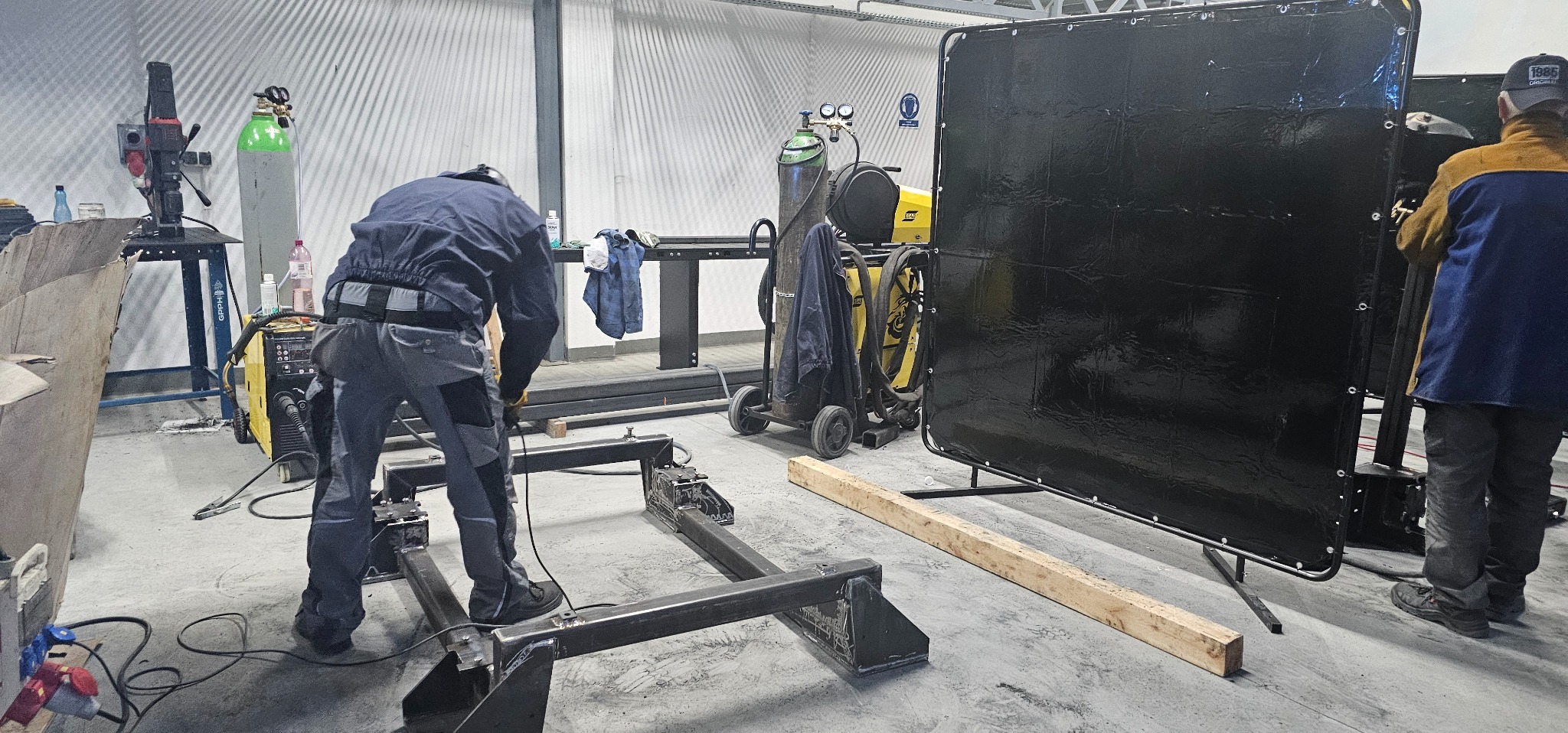 Spawanie konstrukcji stalowej w warsztacie: spawacz w odzieży ochronnej łączy elementy ramy, widoczna spawarka, butla z gazem, parawan spawalniczy i narzędzia.