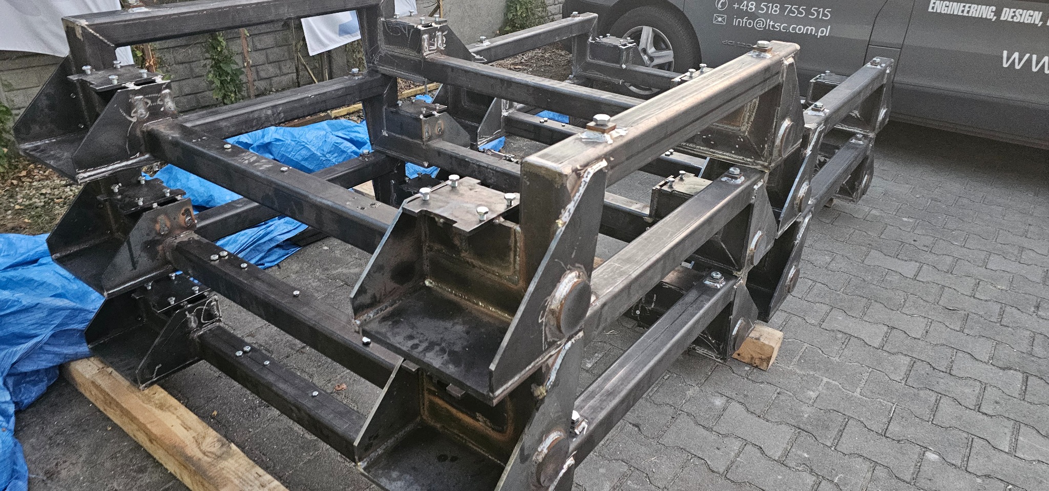 Metalowa konstrukcja spawana z profili stalowych, łączona śrubami, widoczna na zewnątrz budynku, częściowo przykryta niebieską folią.