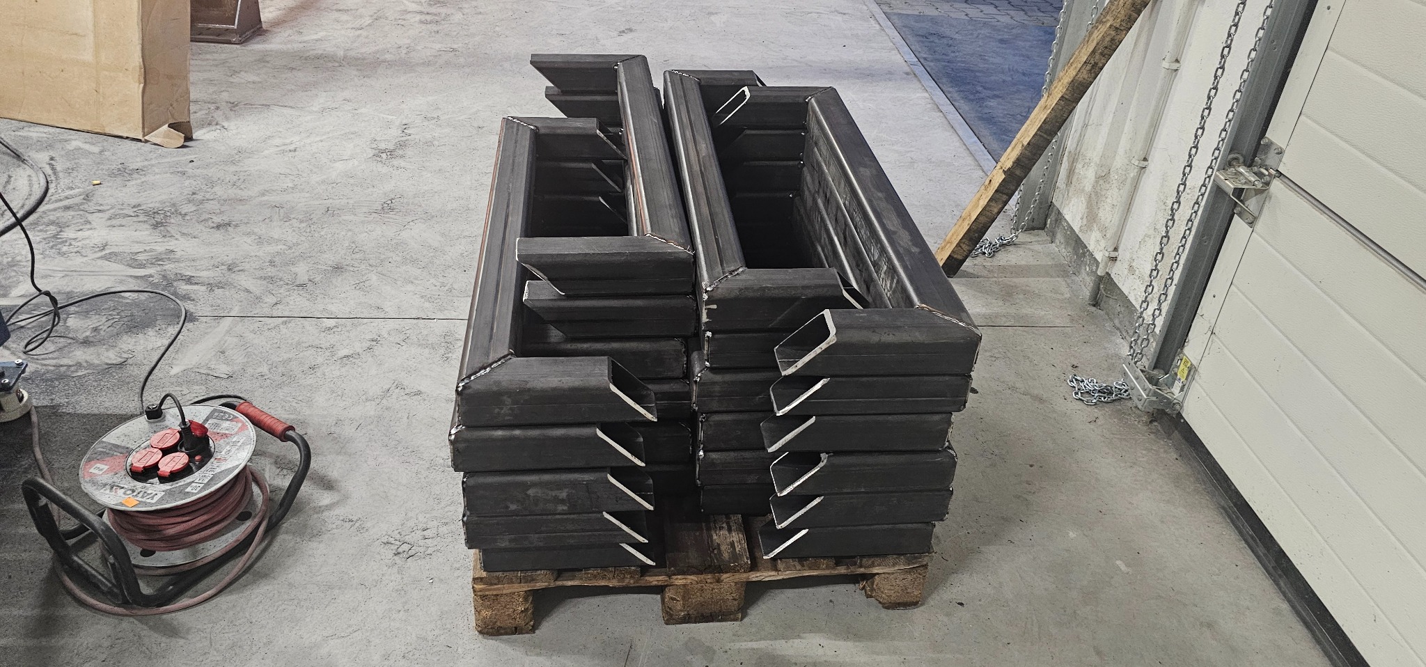 Stos spawanych, metalowych profili zamkniętych o przekroju kwadratowym, ułożonych na palecie w hali produkcyjnej. Widoczny przewód zasilający i brama garażowa w tle.