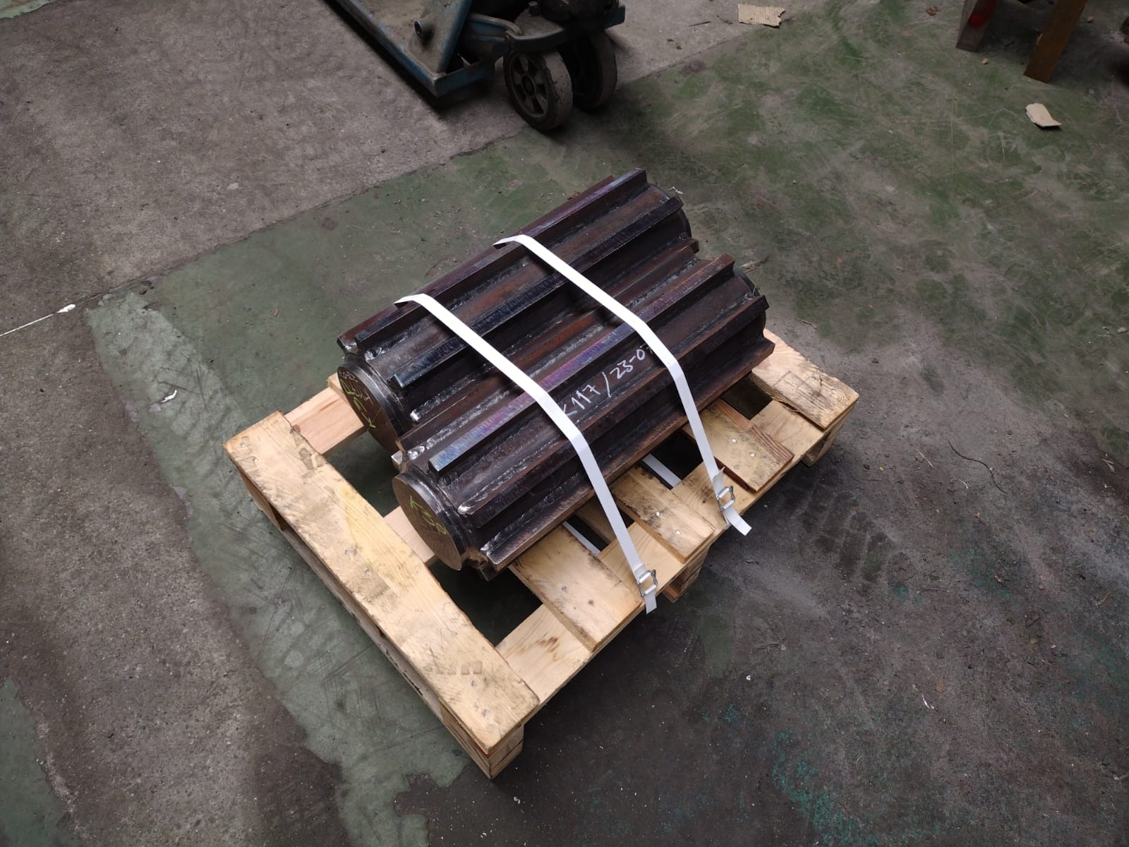 Dwa duże, metalowe koła zębate, zabezpieczone białymi pasami, leżące na drewnianej palecie na tle betonowej posadzki warsztatu. Widoczne ślady obróbki i napisy na powierzchni metalu.