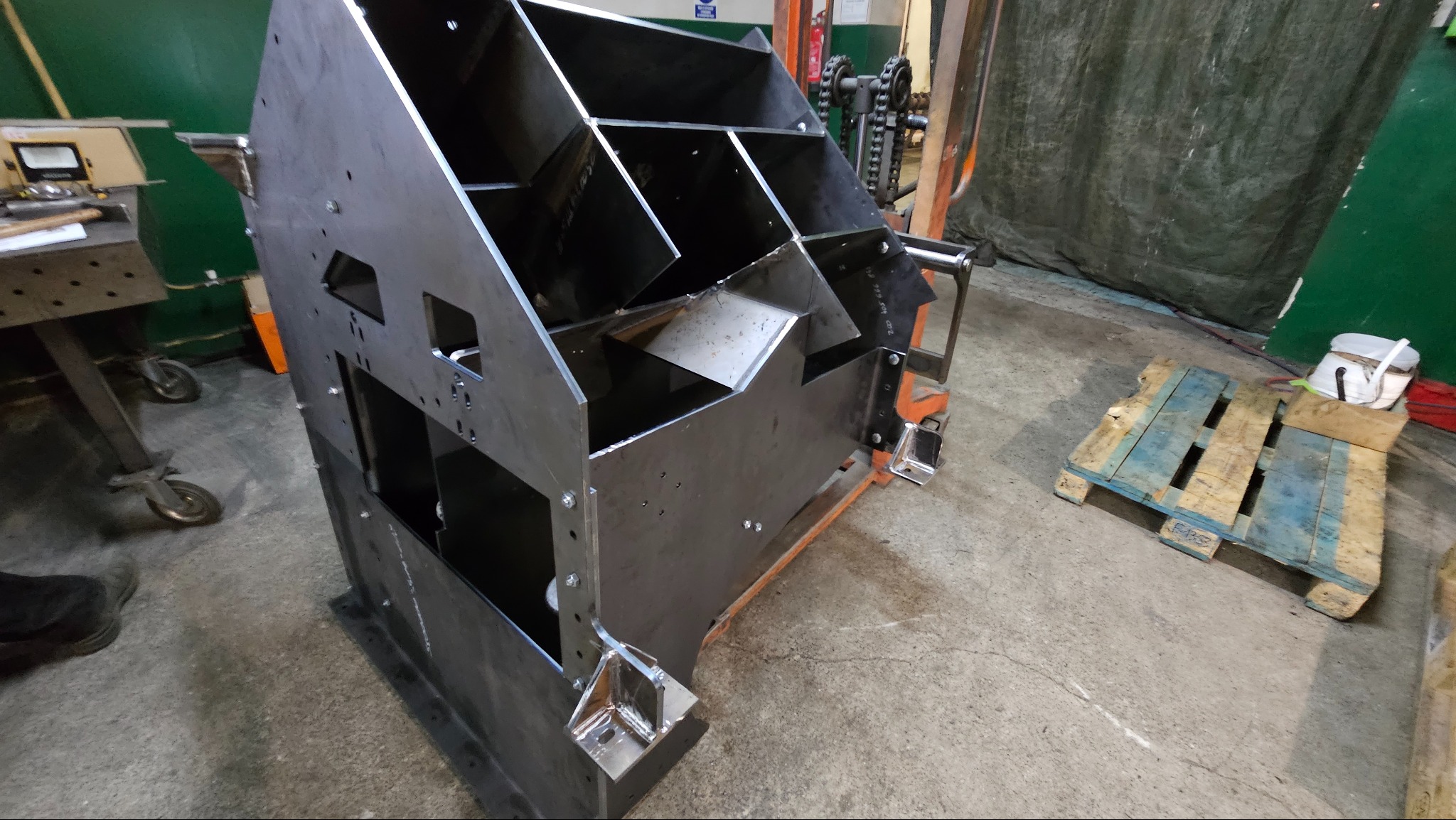 Surowa, stalowa konstrukcja z licznymi przegródkami i otworami, umieszczona na palecie w warsztacie, widoczne ślady spawania i obróbki.