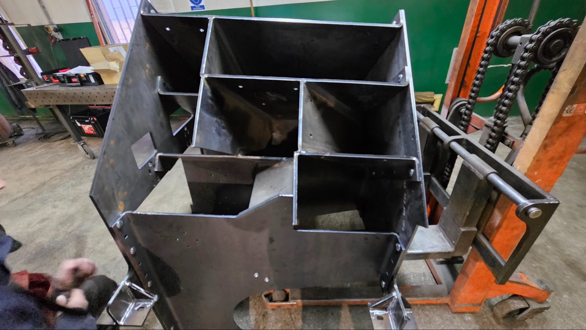 Metalowa konstrukcja z przegródkami, prawdopodobnie regał lub organizer, spawana z blachy i profili, umieszczona na pomarańczowym wózku widłowym w warsztacie.