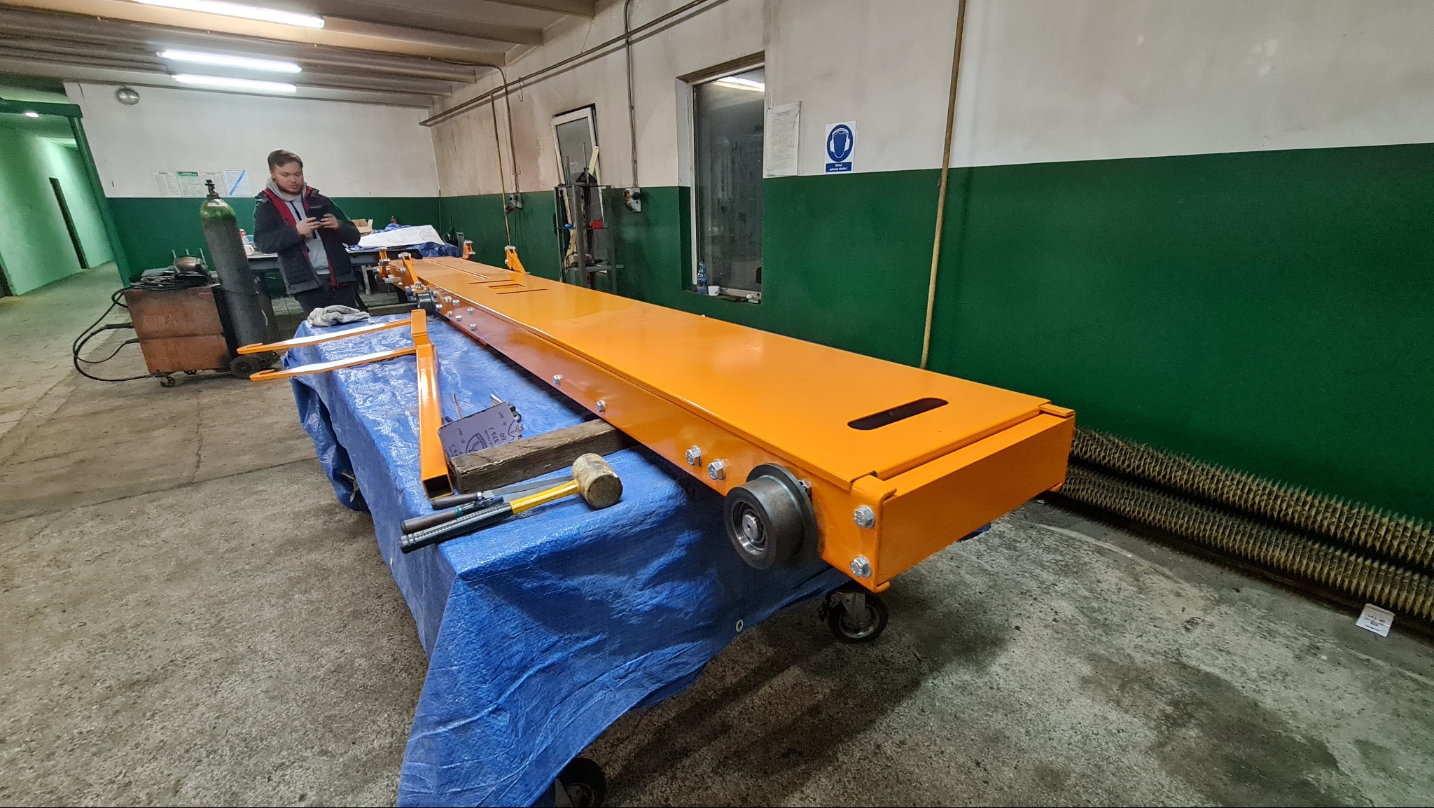 Pomarańczowa konstrukcja stalowa na wózku warsztatowym, widoczne mocowania śrubowe, koła, w tle butla z gazem i pracownik przeglądający telefon w warsztacie.