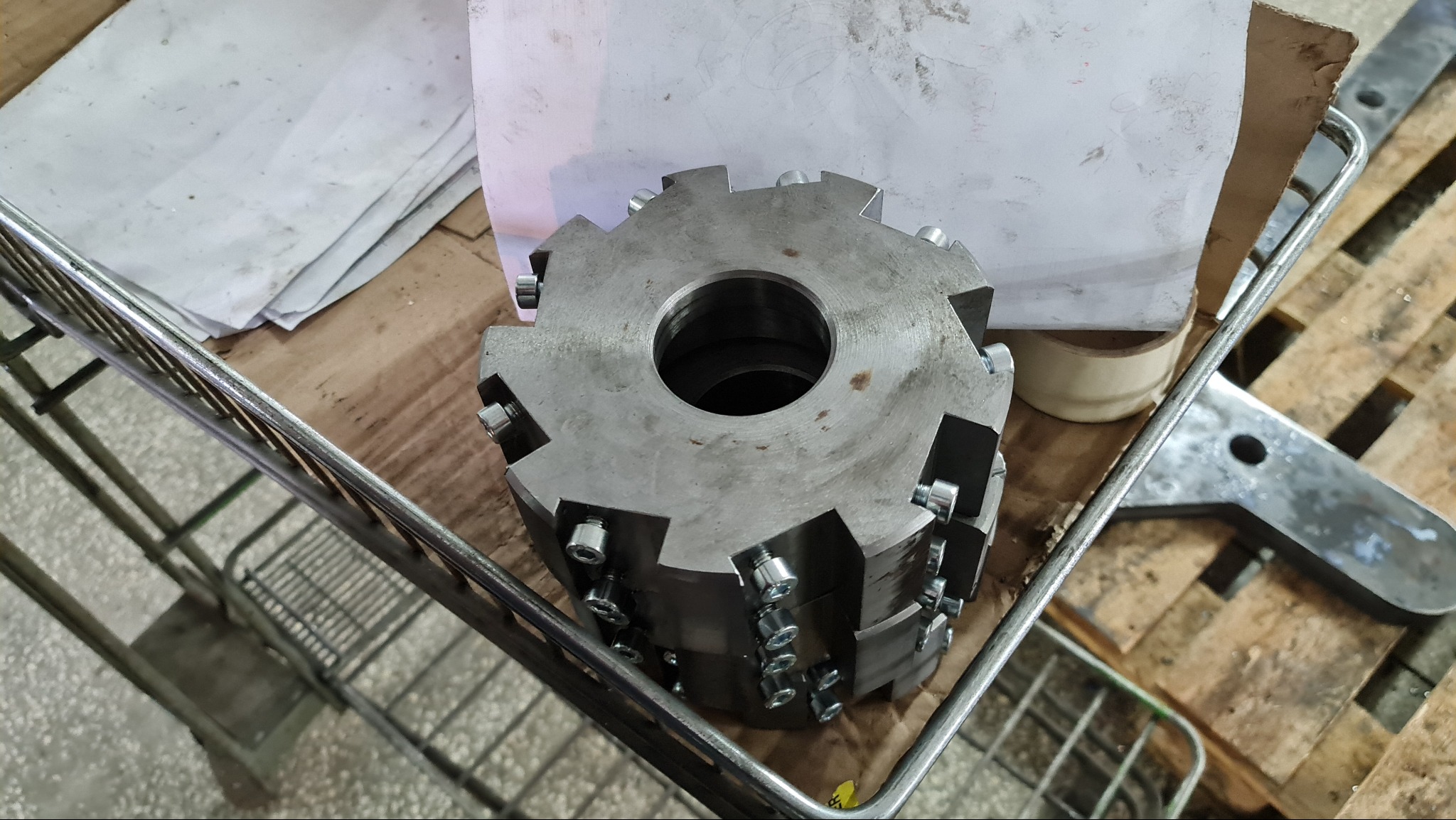 Metalowa głowica frezarska z wymiennymi płytkami skrawającymi, leżąca w metalowym koszu na tle drewnianej palety i papierów.