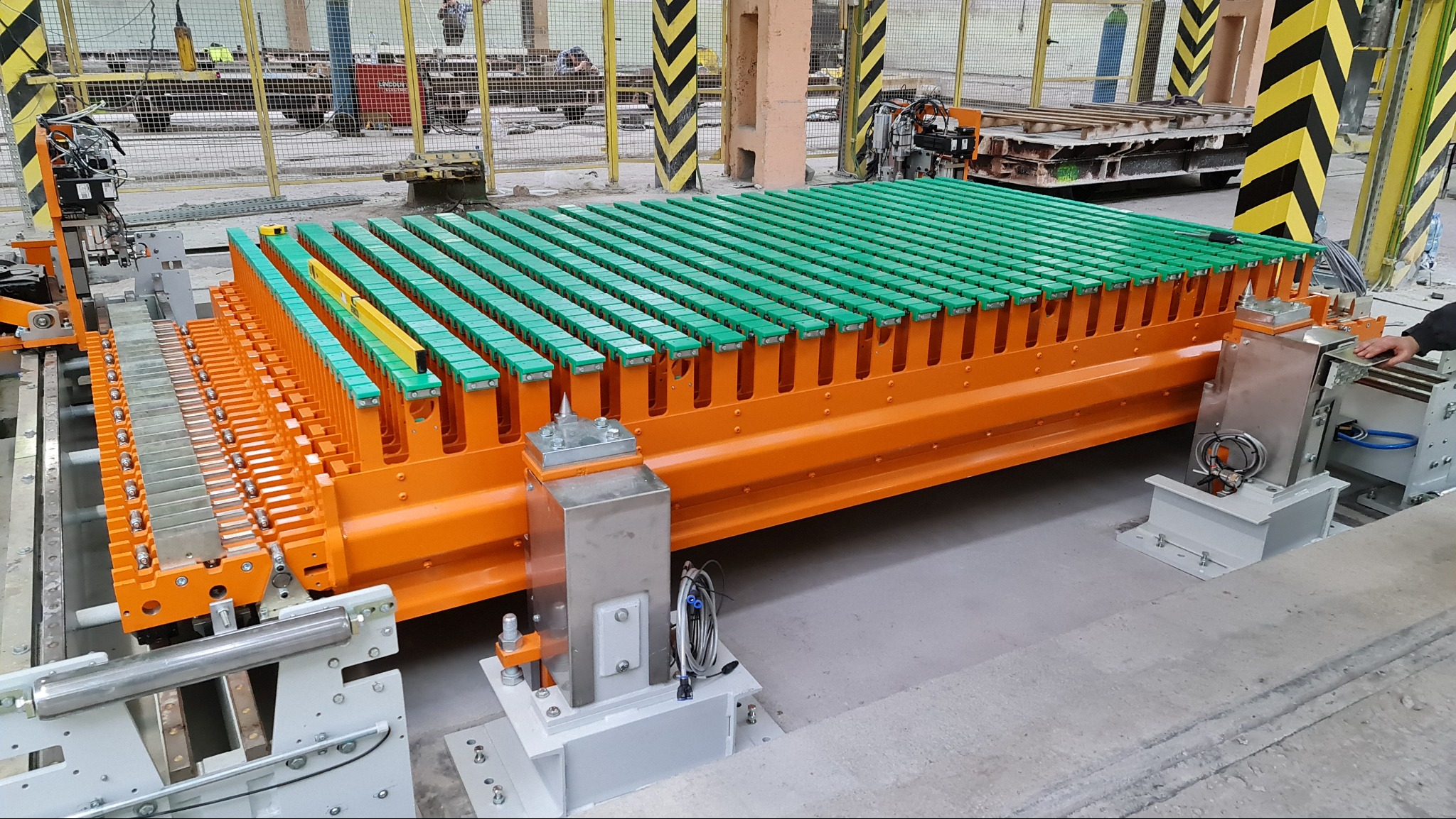 Pomarańczowa konstrukcja z zielonymi elementami, prawdopodobnie część większej maszyny, ustawiona na szarym betonowym podłożu w hali produkcyjnej. Widoczne elementy automatyki i czujniki.