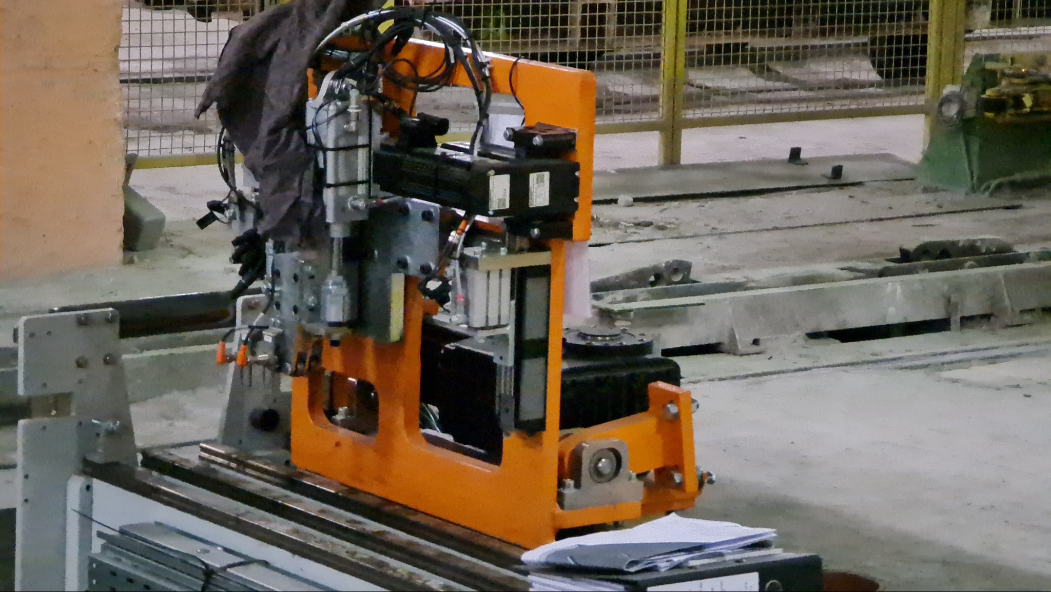 Pomarańczowa frezarka CNC z widocznymi elementami sterującymi i osprzętem, ustawiona w hali produkcyjnej z betonową podłogą i metalowymi barierkami w tle. Na stole leżą dokumenty.