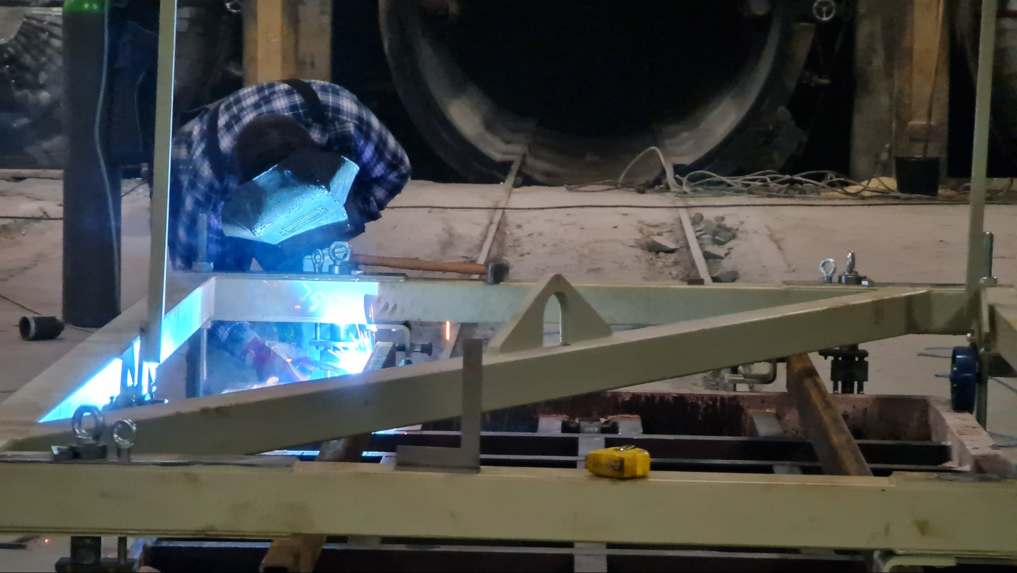 Pracownik w masce spawalniczej łączy metalowe elementy konstrukcji spawarką, widoczne iskry i jarzące się światło łuku elektrycznego, w tle duży, ciemny zbiornik.