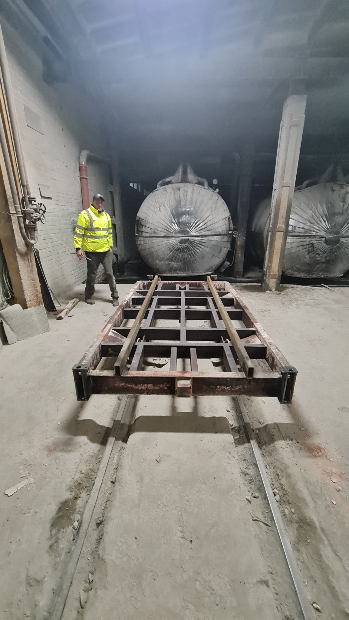 Metalowa rama na szynach w hali produkcyjnej, w tle dwa duże zbiorniki owinięte folią i pracownik w odblaskowej kurtce.