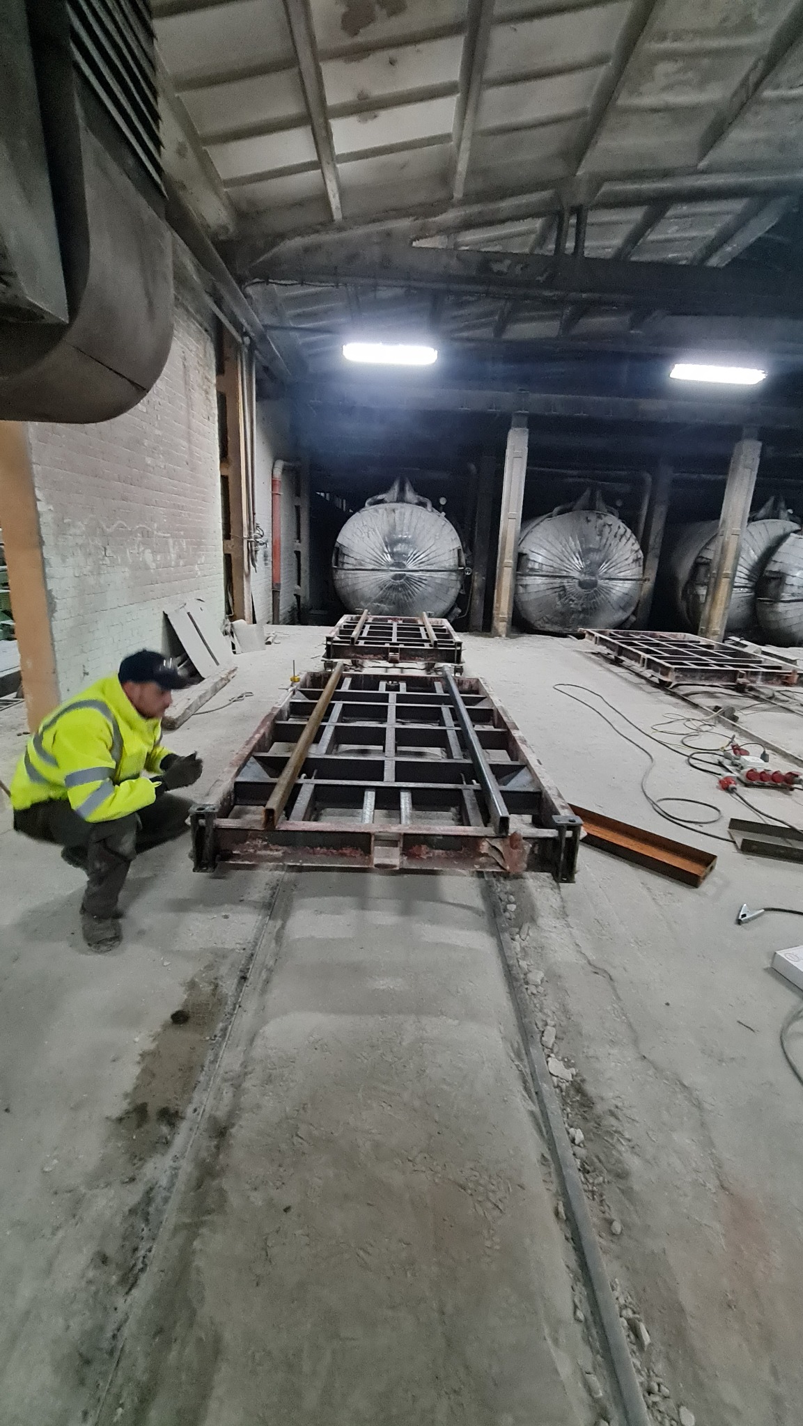 Pracownik w żółtej kamizelce przygotowuje metalową konstrukcję kratową na szynach w hali produkcyjnej, w tle widoczne duże zbiorniki ciśnieniowe.