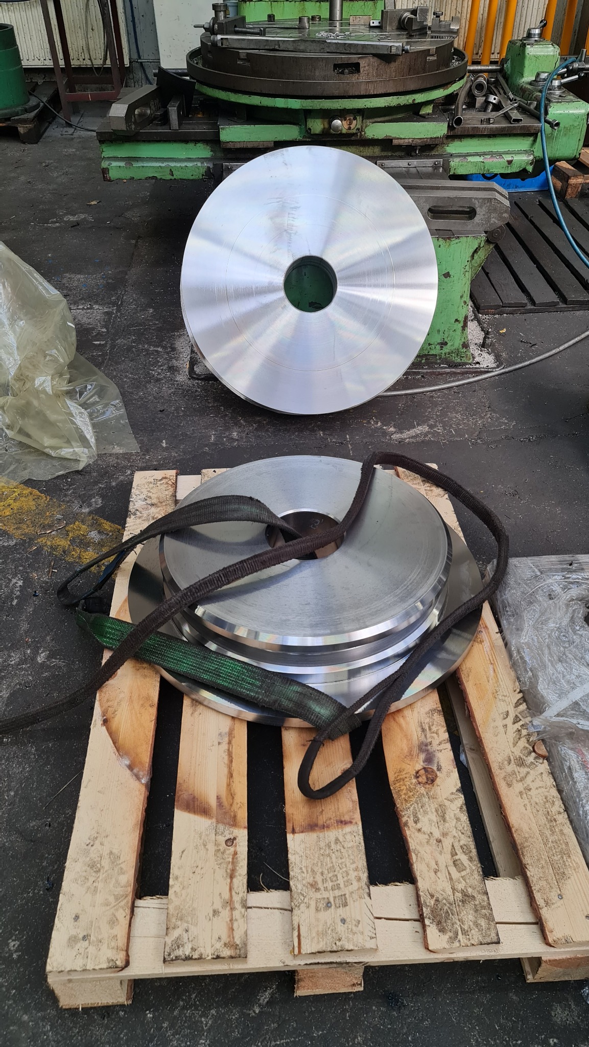 Srebrne, okrągłe elementy z centralnym otworem, ułożone na drewnianej palecie z pasami transportowymi, w tle widoczna zielona tokarka.