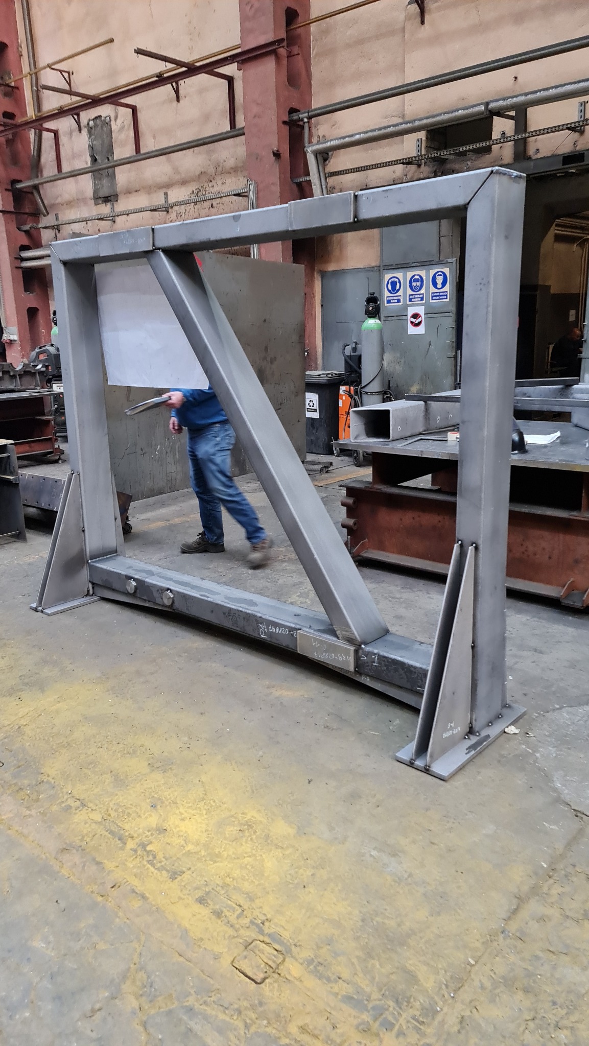 Metalowa rama konstrukcyjna z ukośnym wzmocnieniem, spawana, z widocznymi śladami spawania, ustawiona w hali produkcyjnej, z pracownikiem w tle.