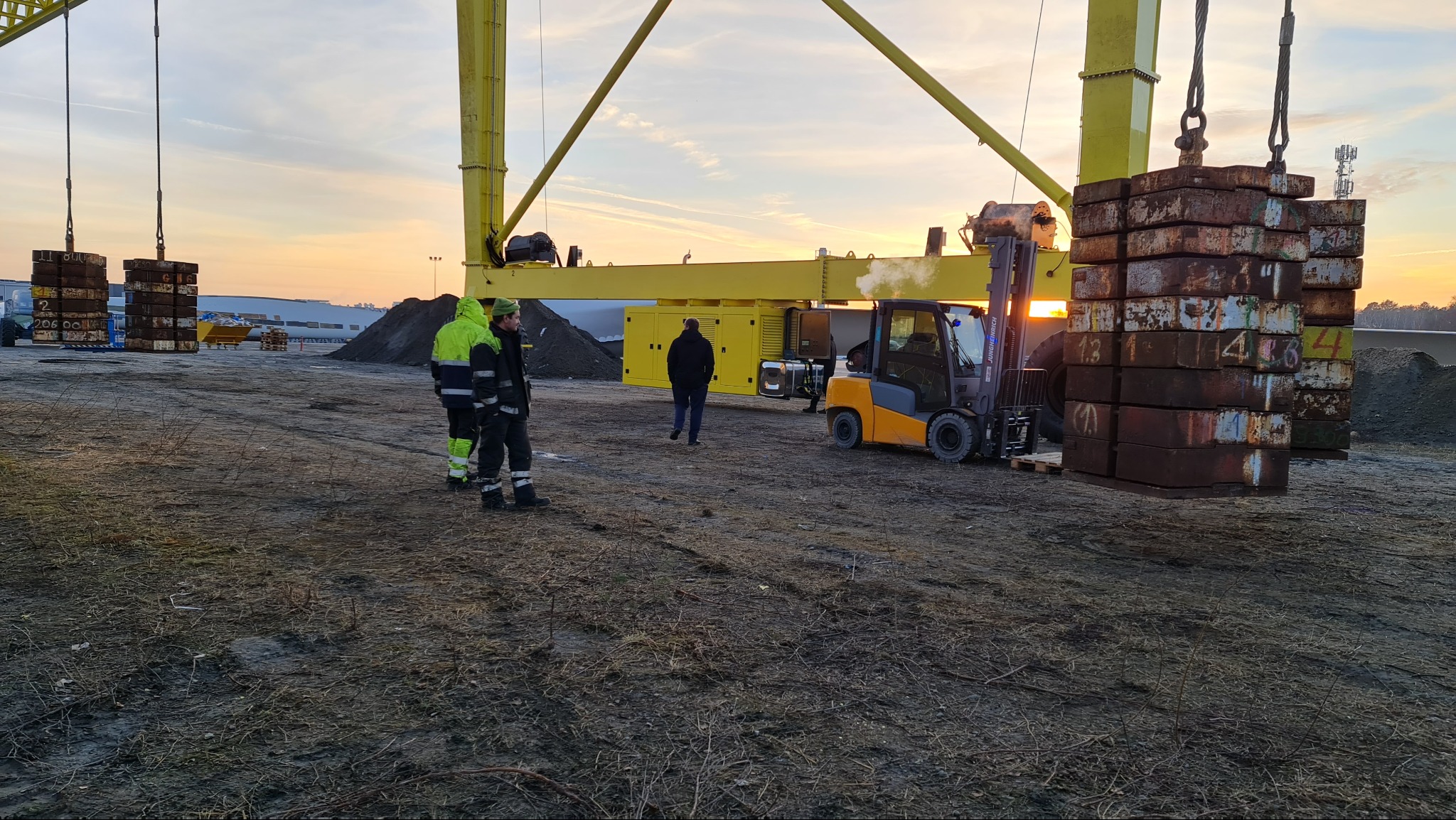 Żółty dźwig podnosi paletę z obciążnikami, w tle wózek widłowy i pracownicy w odblaskowych ubraniach na placu budowy o zachodzie słońca.