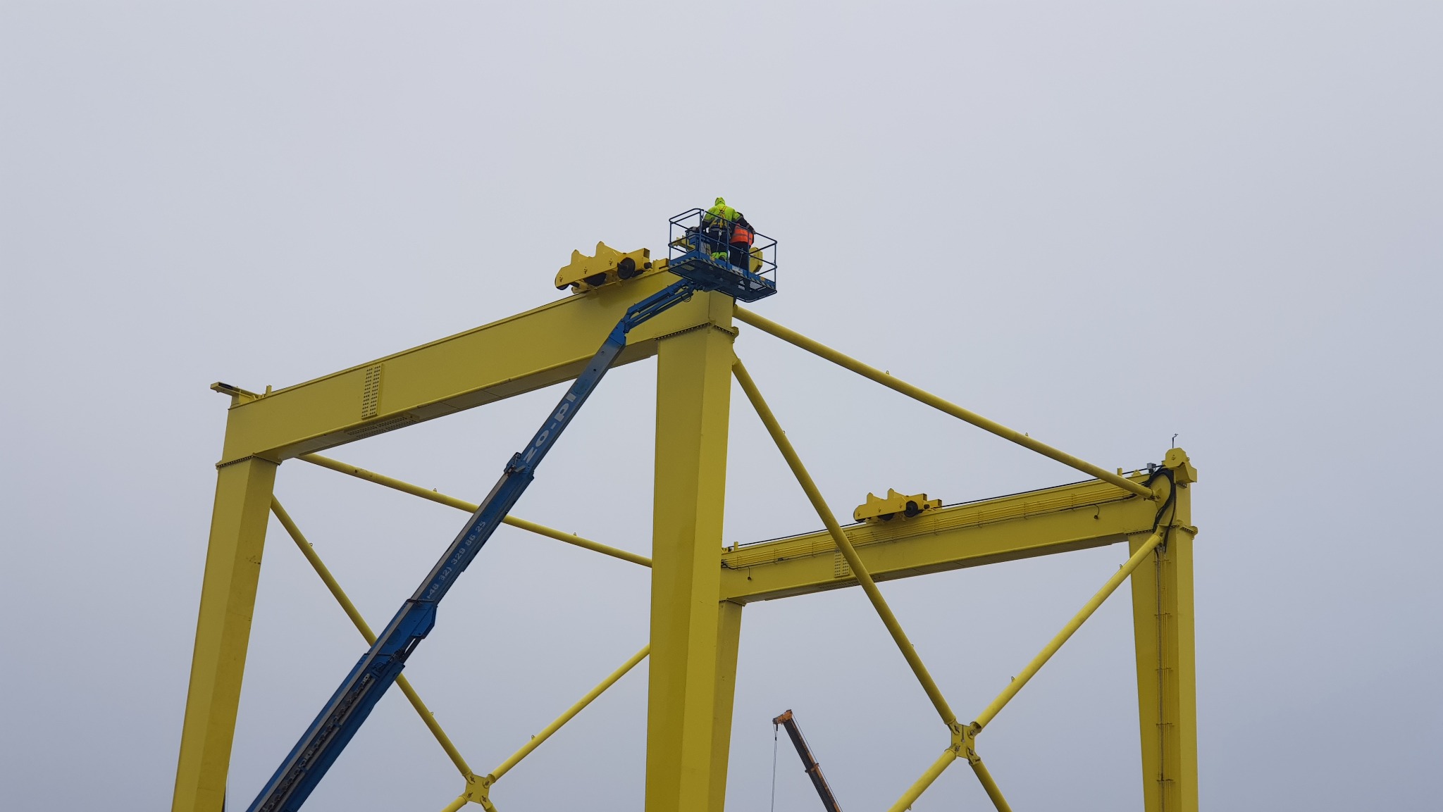 Żółta konstrukcja suwnicy bramowej w trakcie montażu, pracownik na podnośniku koszowym w górnej części konstrukcji, widoczne elementy mocujące i spawane łączenia.