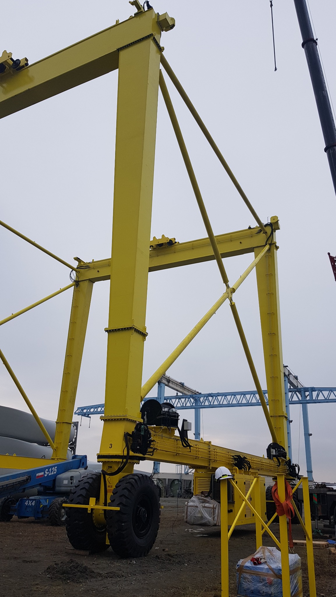 Żółta, mobilna suwnica bramowa na placu budowy, widok od dołu, z widocznymi elementami konstrukcyjnymi, kołami i osprzętem elektrycznym, w tle inne konstrukcje stalowe i podnośnik koszowy.