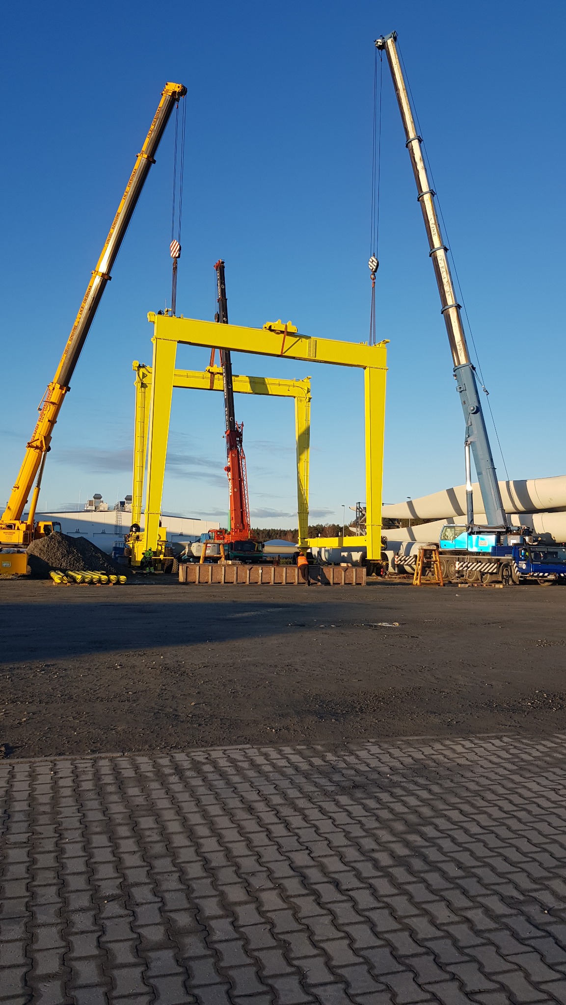 Dwa dźwigi podnoszą żółtą, stalową konstrukcję bramową na placu budowy, w tle widoczne elementy turbin wiatrowych.